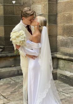 a bride and groom kissing in front of a stone building with white flowers on it