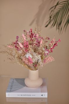 a white vase filled with pink flowers on top of a book next to a plant