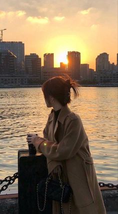 a woman standing next to a body of water holding a black suitcase with the sun setting in the background