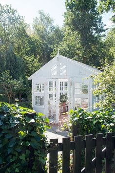 a small white house surrounded by greenery in the middle of a garden with a wooden fence