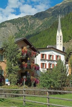 a building with flowers on the outside and a clock tower in the background, surrounded by mountains