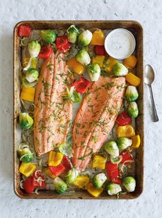 two salmons on a tray with vegetables and dressing next to a fork, knife and spoon