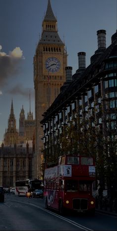 a red double decker bus driving down a street next to tall buildings and a clock tower