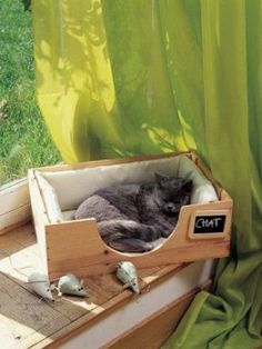 a cat laying in a wooden box on top of a window sill next to a green curtain