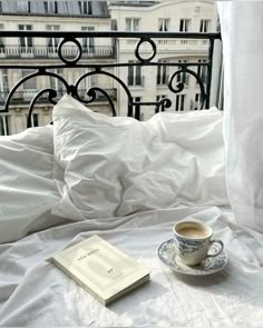 a cup of coffee sitting on top of a white bed next to a book and an open window