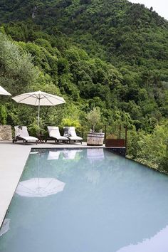 an empty swimming pool with lounge chairs and umbrellas near the edge, surrounded by lush green mountains