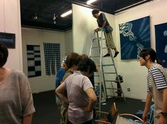 several people are standing around and looking at artwork on the wall, while one man is climbing up a ladder