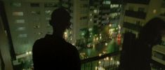 two people standing on a balcony at night looking out over a cityscape with buildings in the background