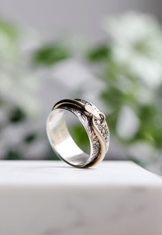 "My Snake In The Grass ring features a 10mm (3/8\") wide floral inner band and an outer ring in the shape of an ouroboros (a snake eating its own tail) that spins freely around the inner band. The snake sneaks around the foliage of the wide band, held in place by the flared ends of that band. These flared ends give the ring a comfortable fit that glides onto your finger. This ring is available in sterling silver with either a bronze, sterling silver, or 14k yellow gold snake. Also available with Engraved Snake Ring For Anniversary, Ouroboros Ring, Snake Eating, Snake In The Grass, Silver Mens Ring, Womens Ring, Sterling Silver Mens Rings, Silver Spinner Rings, Snake Ring