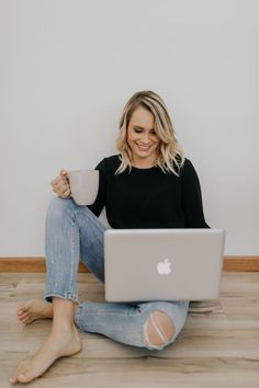 a woman sitting on the floor holding a coffee cup and looking at her laptop computer