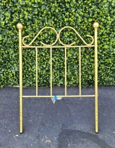 a gold metal headboard with two posts and a blue ball at the top, in front of a green hedge