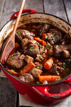 a red pot filled with meat and carrots on top of a wooden table next to a spoon