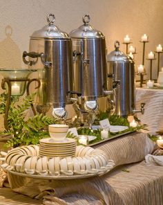 a table topped with lots of silver pots and pans covered in greenery next to candles