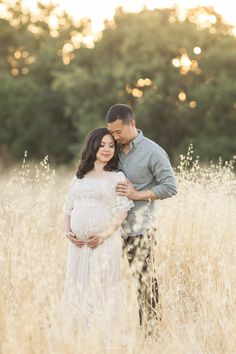 a pregnant couple standing in tall grass with the sun shining down on them and holding each other