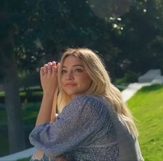 a beautiful blonde woman sitting on top of a green grass covered park bench next to a tree