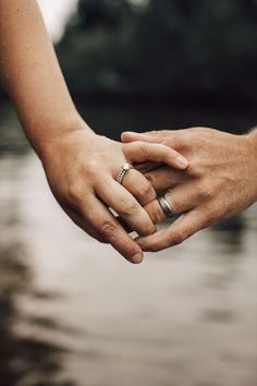 two people holding hands over water with trees in the background and one person wearing a wedding ring