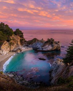 the beach is surrounded by large rocks and trees at sunset with pink clouds in the sky