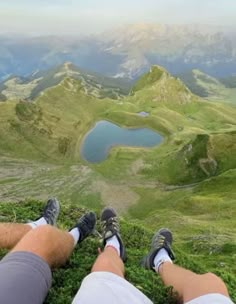 two people sitting on top of a lush green hillside with mountains in the back ground