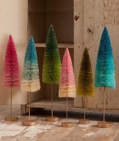 five small trees are lined up in different colors on the floor next to a wooden shelf