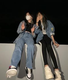 two young women sitting on the edge of a wall with their faces covered by masks