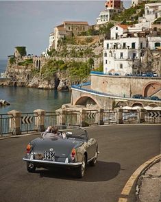 an old car is driving down the road by the water in front of some buildings