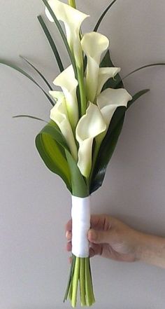 a hand holding a bouquet of white calla lilies and greenery in front of a wall