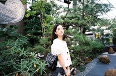 a woman standing in the middle of a garden holding a black purse and looking up