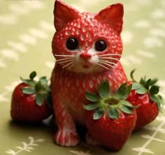 a red cat figurine sitting next to three strawberries on a green tablecloth