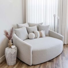 a round couch with pillows on it next to a vase and lamp in front of a window