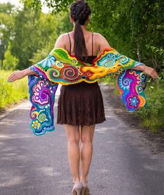 a woman in a dress walking down a road with her arms spread open and colorful shawl draped over her shoulders