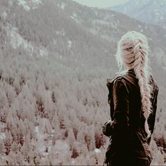 a woman with long blonde hair standing in front of a snowy mountain range looking at the trees