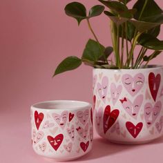 two cups with hearts painted on them are next to a potted plant and pink background