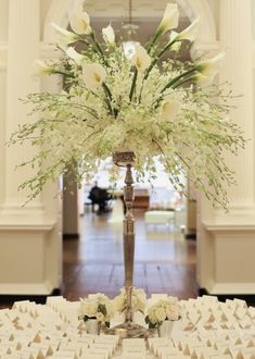 a tall vase with white flowers on top of a table covered in place settings for seating