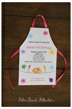 an apron shaped birthday party card on a wooden table with pink ribbon and paint splatters