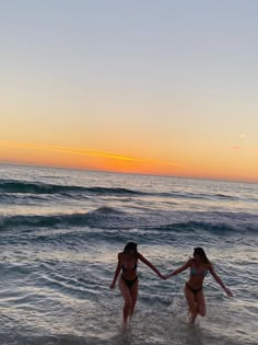 two women in bikinis holding hands while walking into the ocean at sunset or dawn