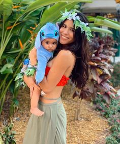 a woman holding a baby in her arms and smiling at the camera while standing next to plants