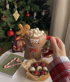 a person holding a mug filled with marshmallows in front of a christmas tree