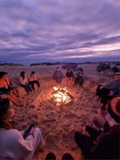 a group of people sitting around a fire pit on the beach at sunset or dawn