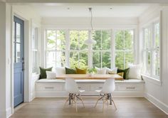 a dining room table with white chairs and green pillows in front of the window seat