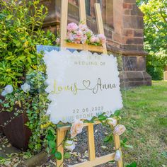 a wedding sign with flowers on it in front of a brick wall and shrubbery