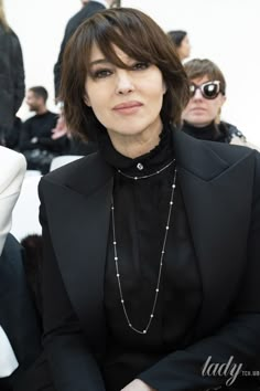 a woman sitting in front of a group of people wearing black clothing and pearls on her necklace