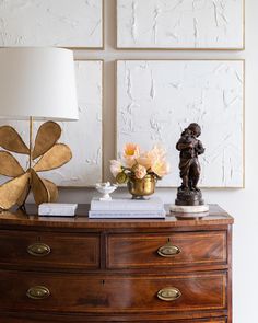 a wooden dresser topped with a lamp next to two paintings and a vase filled with flowers