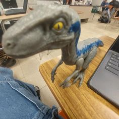 a toy dinosaur is sitting on a desk next to a laptop computer and people in the background