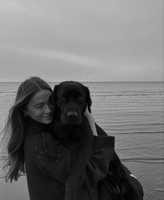 a woman holding a black dog on the beach near the ocean with her eyes closed