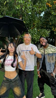 two men and a woman standing under an umbrella in the grass with one giving thumbs up