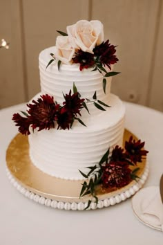 a white wedding cake with red flowers and greenery on the top, sitting on a gold plate