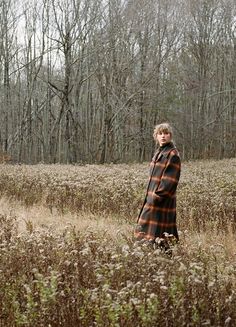 a woman standing in the middle of a field with tall grass and trees behind her