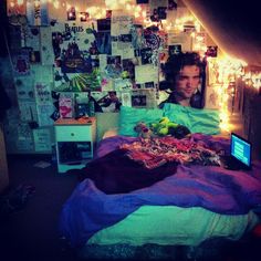 a man sitting in bed with his laptop computer on the table and lights strung up behind him