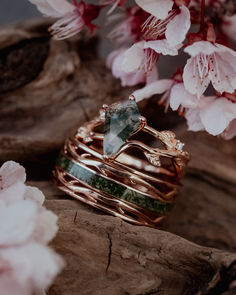 A matching wedding ring set from Staghead Designs sits on a piece of wood surrounded by cherry blossoms. On top sits the "Artemis On The Vine" engagement ring with a branch/leafy shank with diamond accents & a kite cut moss agate center stone. Below sits our men's "Artemis" wedding band with antler/branch cutout details & a centered channel with moss inlay. These rings are shown in 14K rose gold. Masculine Moss Agate Ring, Mens Wedding Rings Moss Agate, Mens Moss Agate Ring, Men’s Wedding Rings Moss Agate, Whiskey Barrel Wedding Ring, Luxury Nature-inspired Rings With Moss Agate, Matching Wedding Ring Sets, Antler Wedding Rings, Matching Wedding Ring