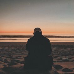 a man sitting on top of a sandy beach next to the ocean under a sunset